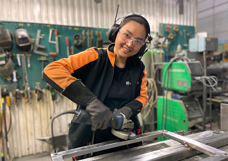Mörkhårig yngre kvinna står i en verkstad och arbetar. Bakom henne hänger olika verktyg och det står maskiner på golvet.