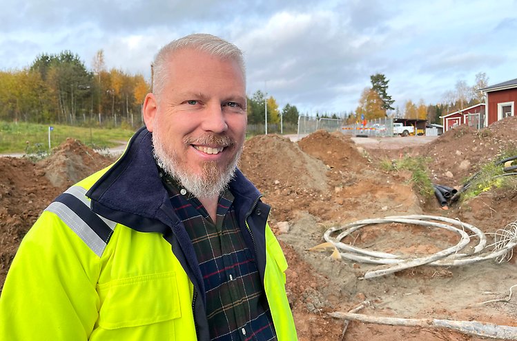 Glad man i varseljacka och skjorta står på arbetsplats med hus i bakgrunden.