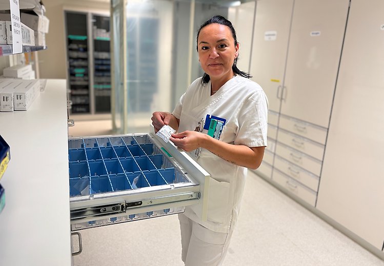 Kvinna i vita vårdkläder står i någon form av laboratorium. Hon placerar saker i fack och tittar in i kameran.